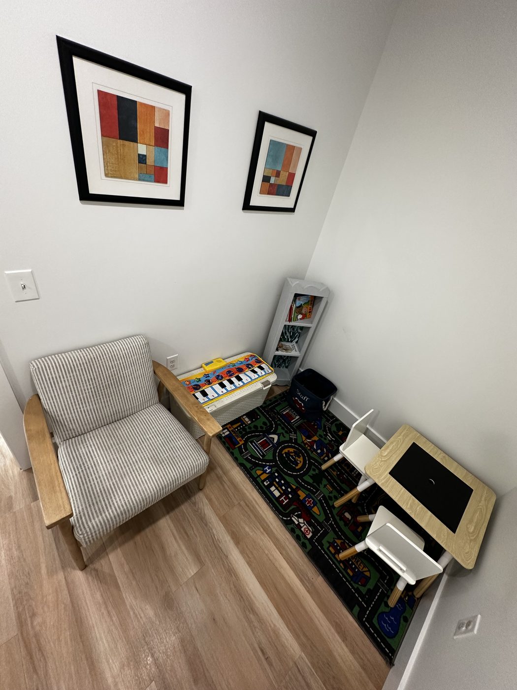 A corner set up for children, with a patterned play rug, a small upholstered chair, a low bookshelf of games and framed prints on the wall